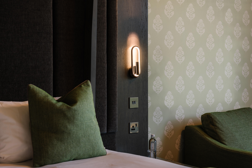 Close-up of a hotel room bed with green cushions, a wall light, power outlets, and patterned wallpaper.
