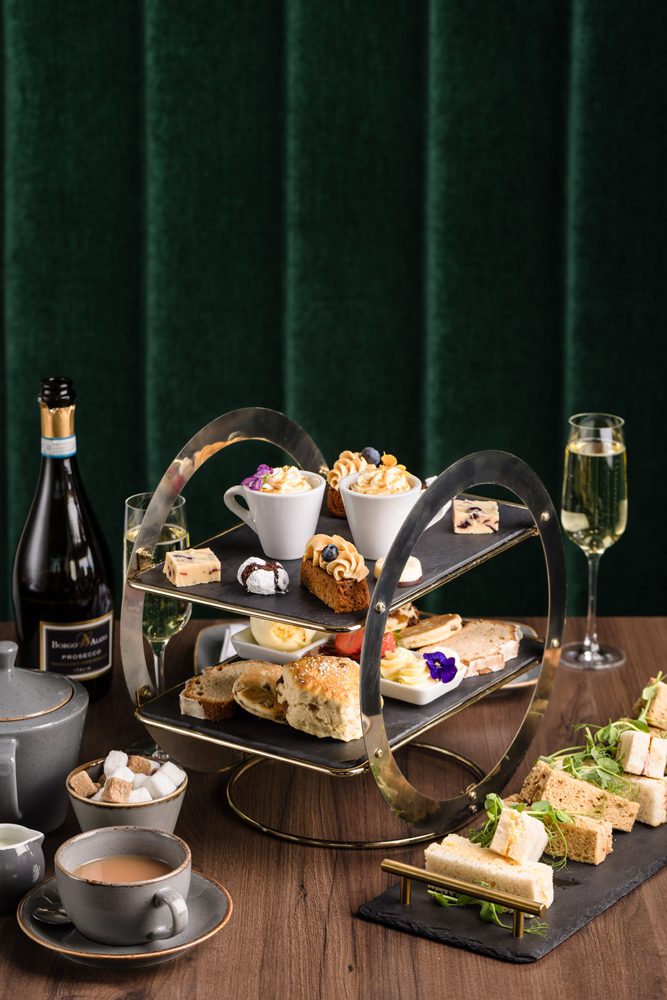 A tiered tray with assorted pastries, sandwiches, tea, and champagne sits on a wooden table with a dark green backdrop.