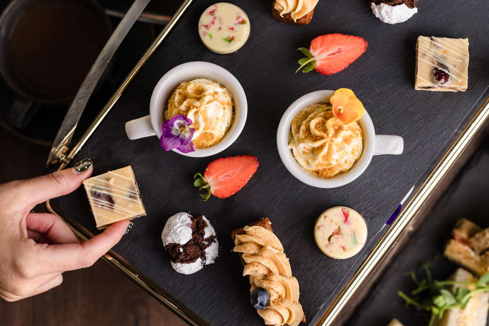 An assortment of pastries, chocolates, and fruit arranged on a slate tray, with a hand reaching for a square treat.