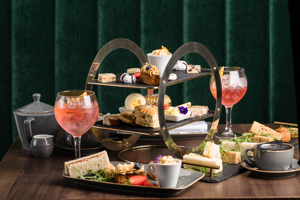 A tiered tray with assorted pastries, scones, and sandwiches, plus tea, pink drinks, and a teapot on a wooden table.