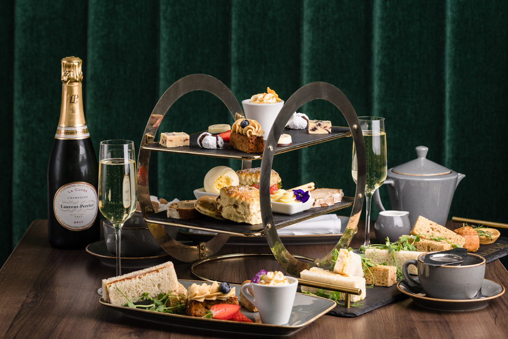 A three-tiered afternoon tea stand with assorted pastries, sandwiches, scones, tea, and a bottle of champagne.