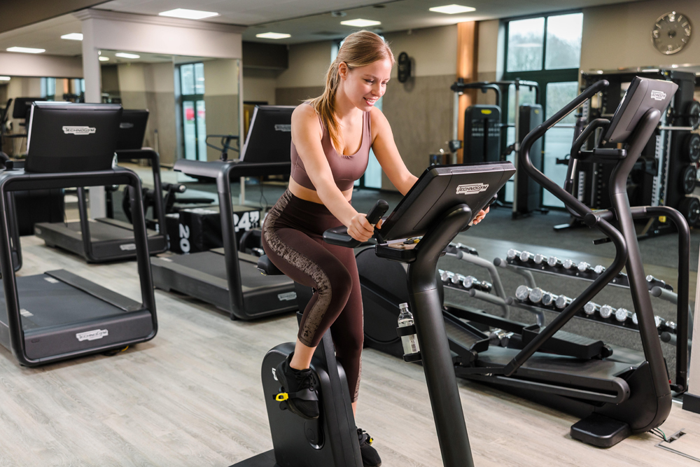 Woman exercising on a stationary bike in a gym, surrounded by weights and other fitness equipment.