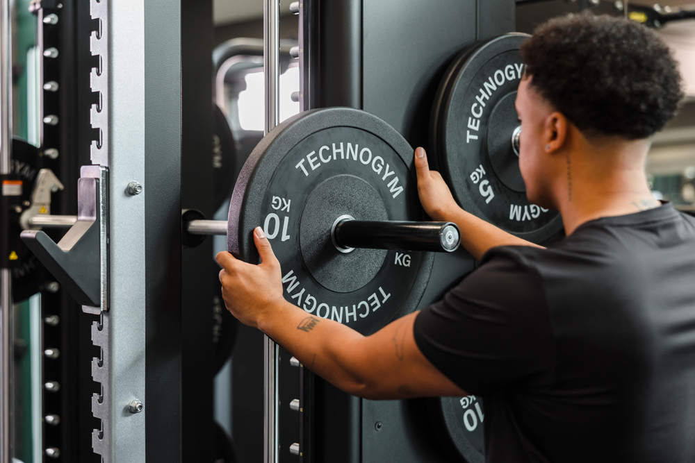 Person placing a 10 kg weight plate onto a barbell in a gym setting.