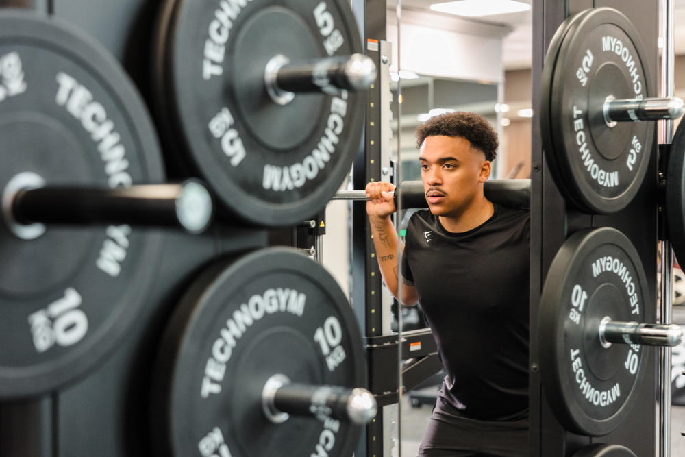 A person wearing a black shirt performs a barbell squat in a gym with weight plates labeled 