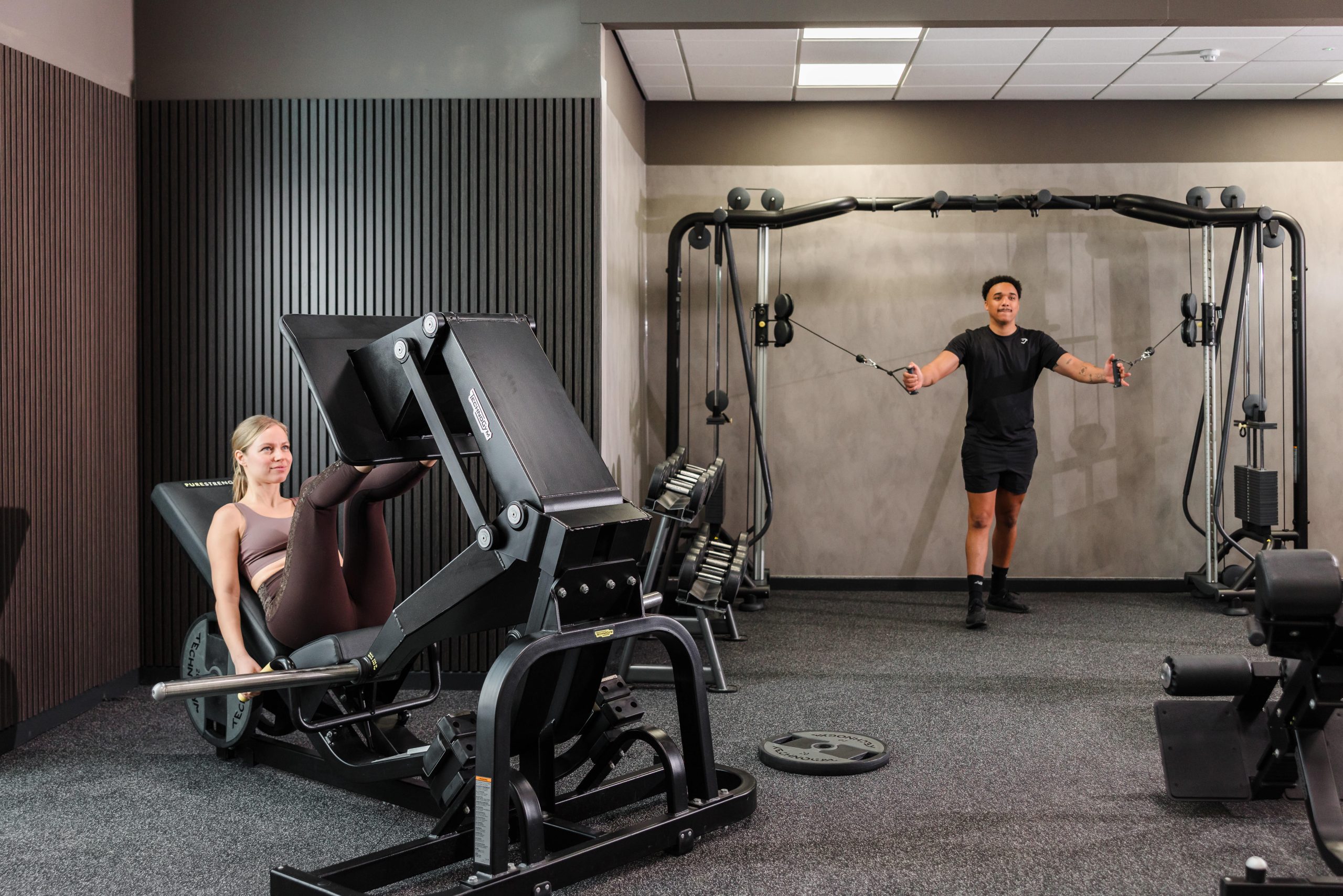 A woman uses a leg press machine while a man does cable exercises in the refurbished Vale Resort South Wales Gym.