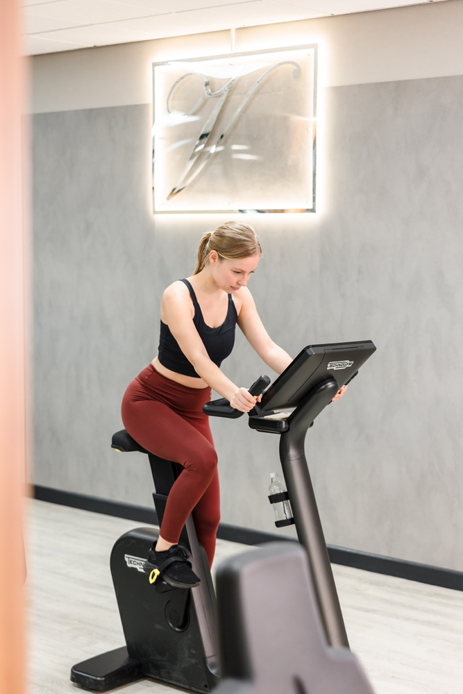 A woman in workout clothes uses a stationary exercise bike in a modern Hotel Gym South Wales with a gray wall and sign behind her.