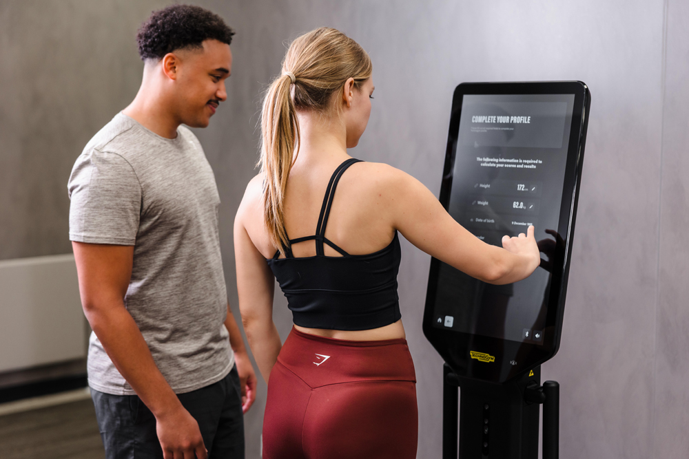 A woman uses a touchscreen fitness machine while a man stands by her side in a gym with a creche.