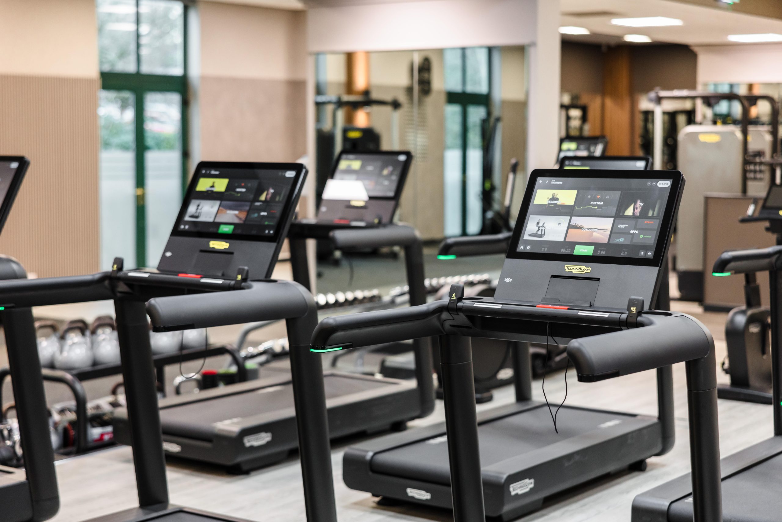 Several modern treadmills line the bright, refurbished Vale Resort South Wales Gym with digital screens and large windows.