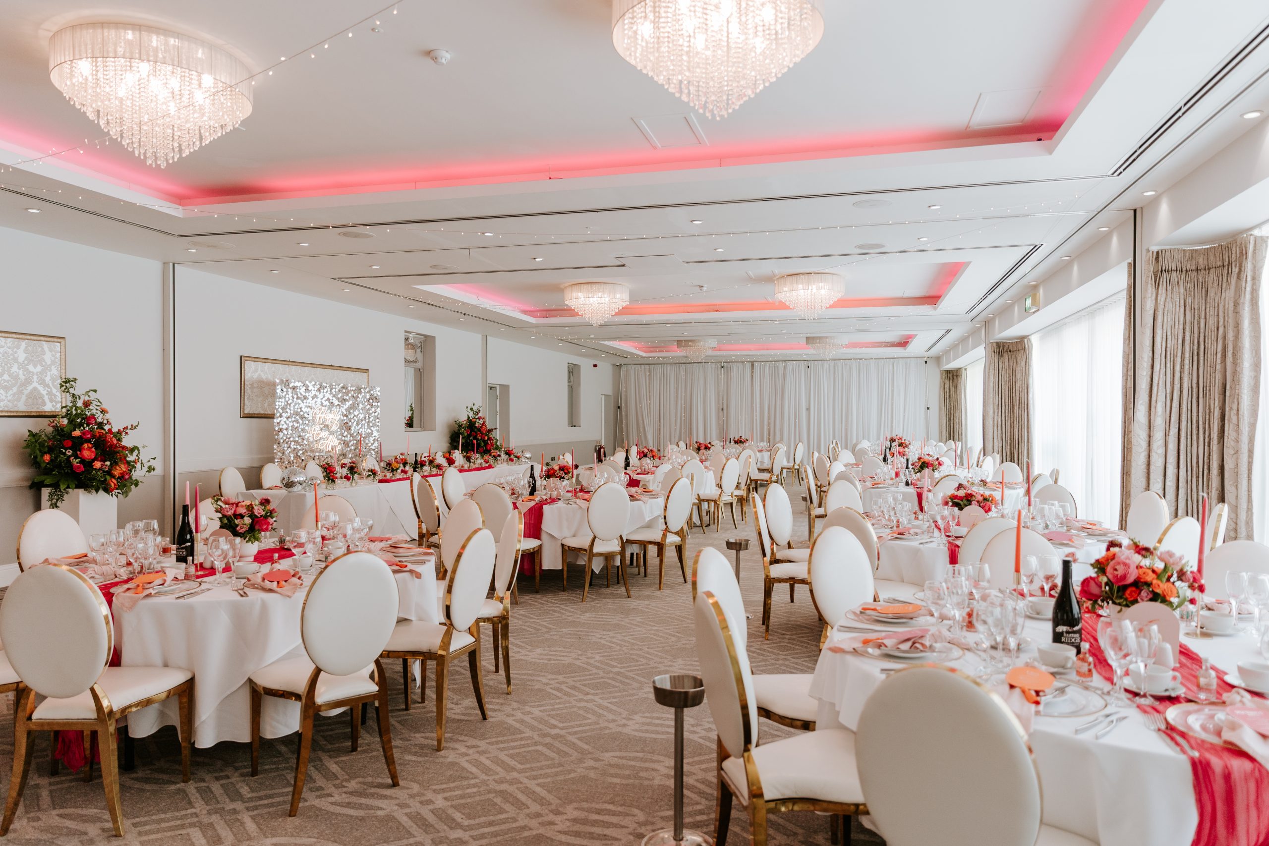 A banquet hall with round tables set for an event, decorated with pink accents and floral centerpieces.