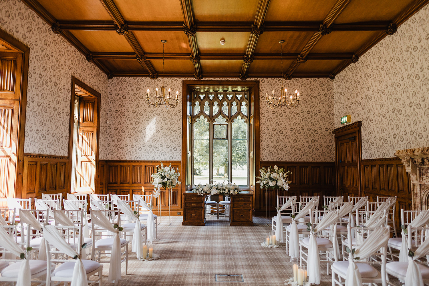 Elegant indoor wedding ceremony setup with white chairs, floral arrangements, and large ornate window in a wood-paneled room.