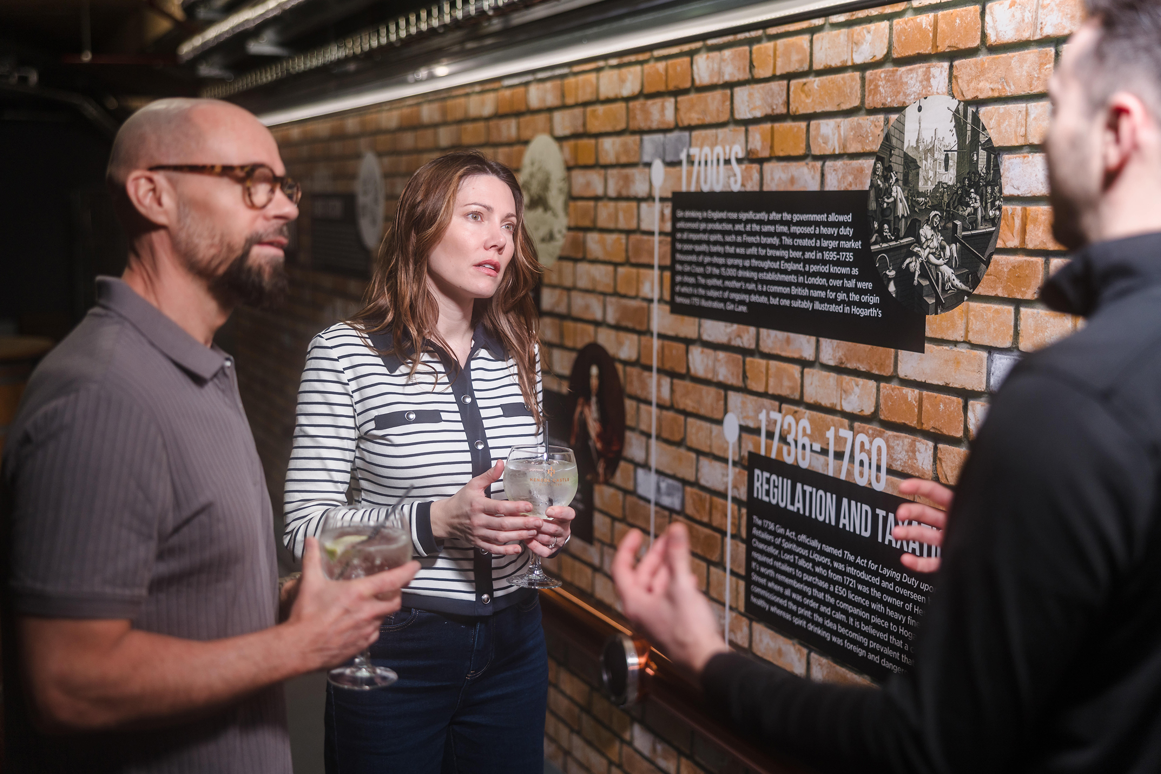 Three adults stand indoors at the estate, holding drinks and reading historical information on a brick wall.
