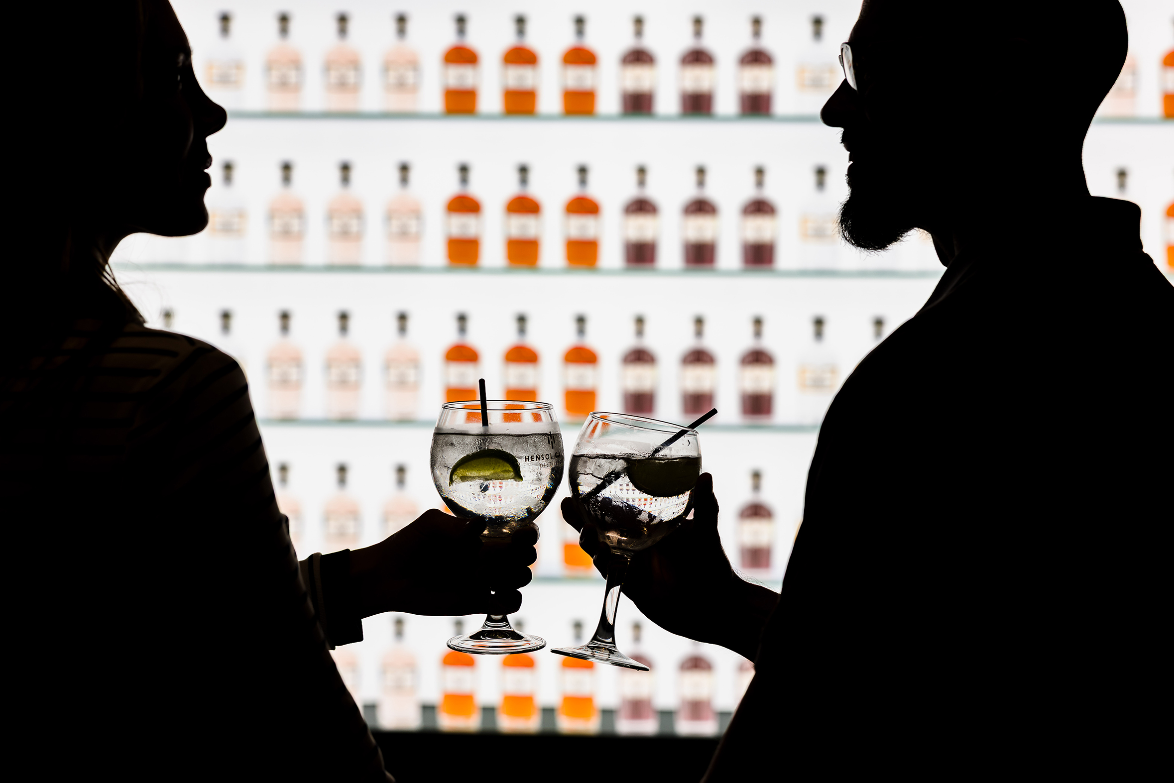 Silhouettes of two people clinking cocktails during stylish hotel breaks South Wales, bottles lined up on a bright shelf.