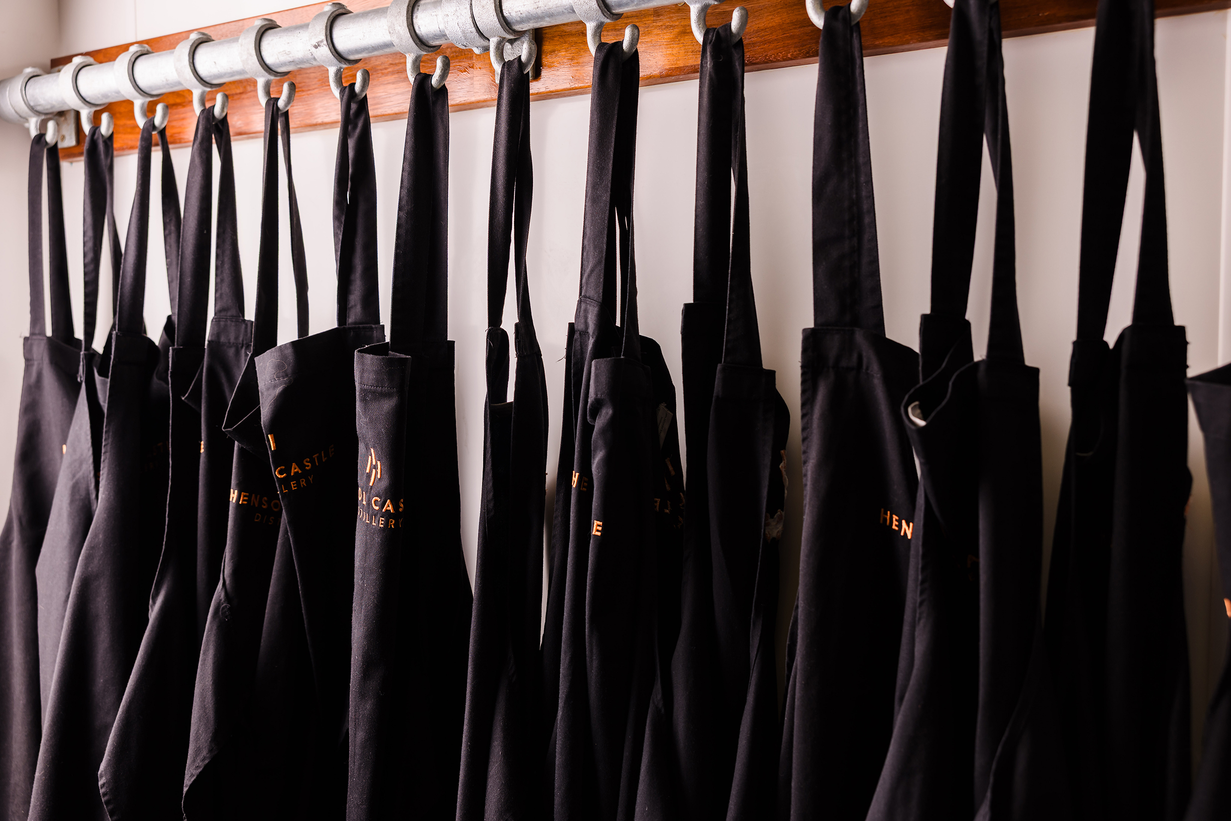 Black aprons for team building hang neatly on metal hooks attached to a wooden rack against a white wall.