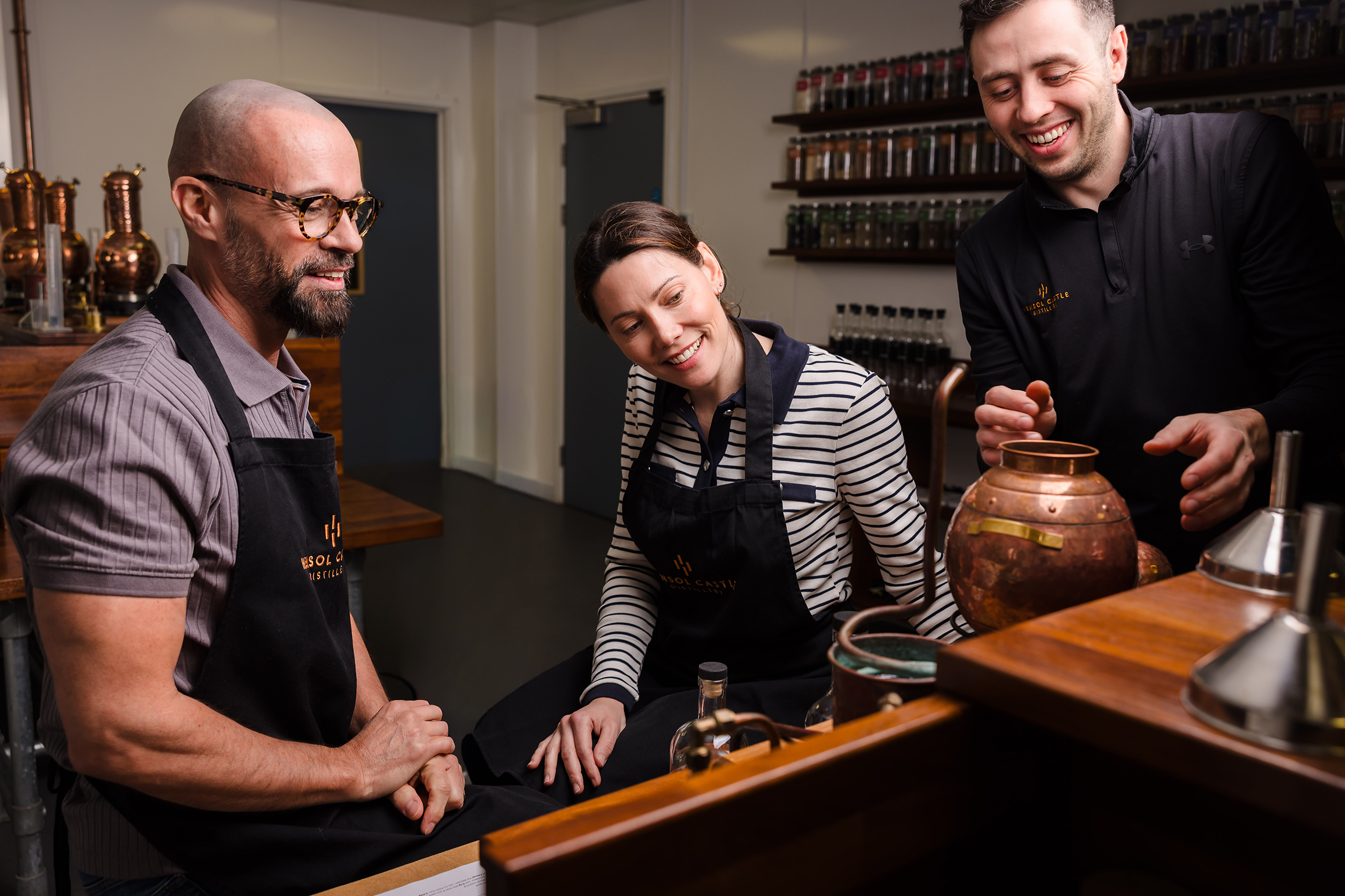 Three people in aprons smile and work together with distilling equipment, discussing hotel room bookings in a modern kitchen.