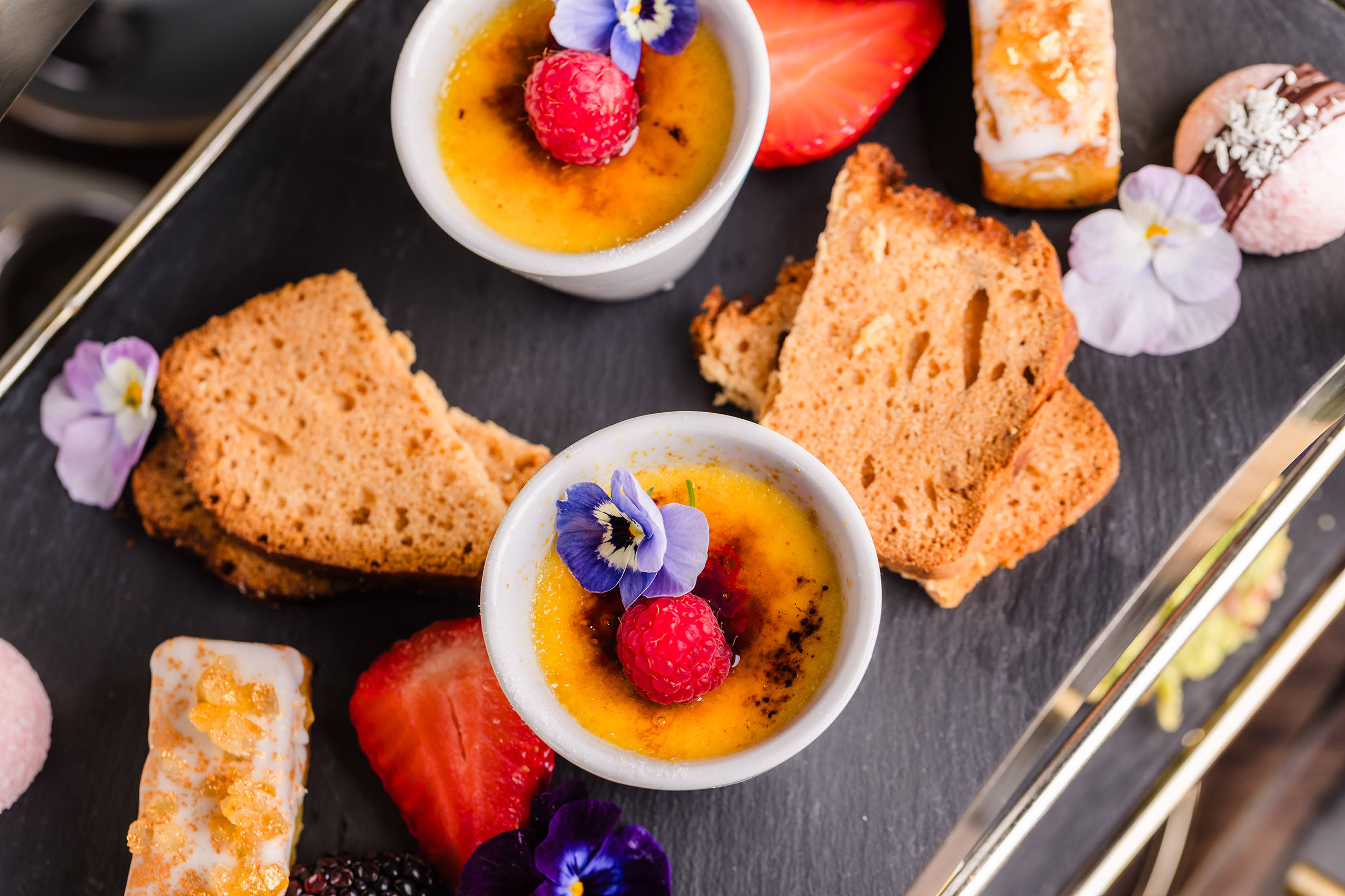 Two ramekins of crème brûlée for afternoon tea, topped with berries, edible flowers, toast, fruit, and pastries.