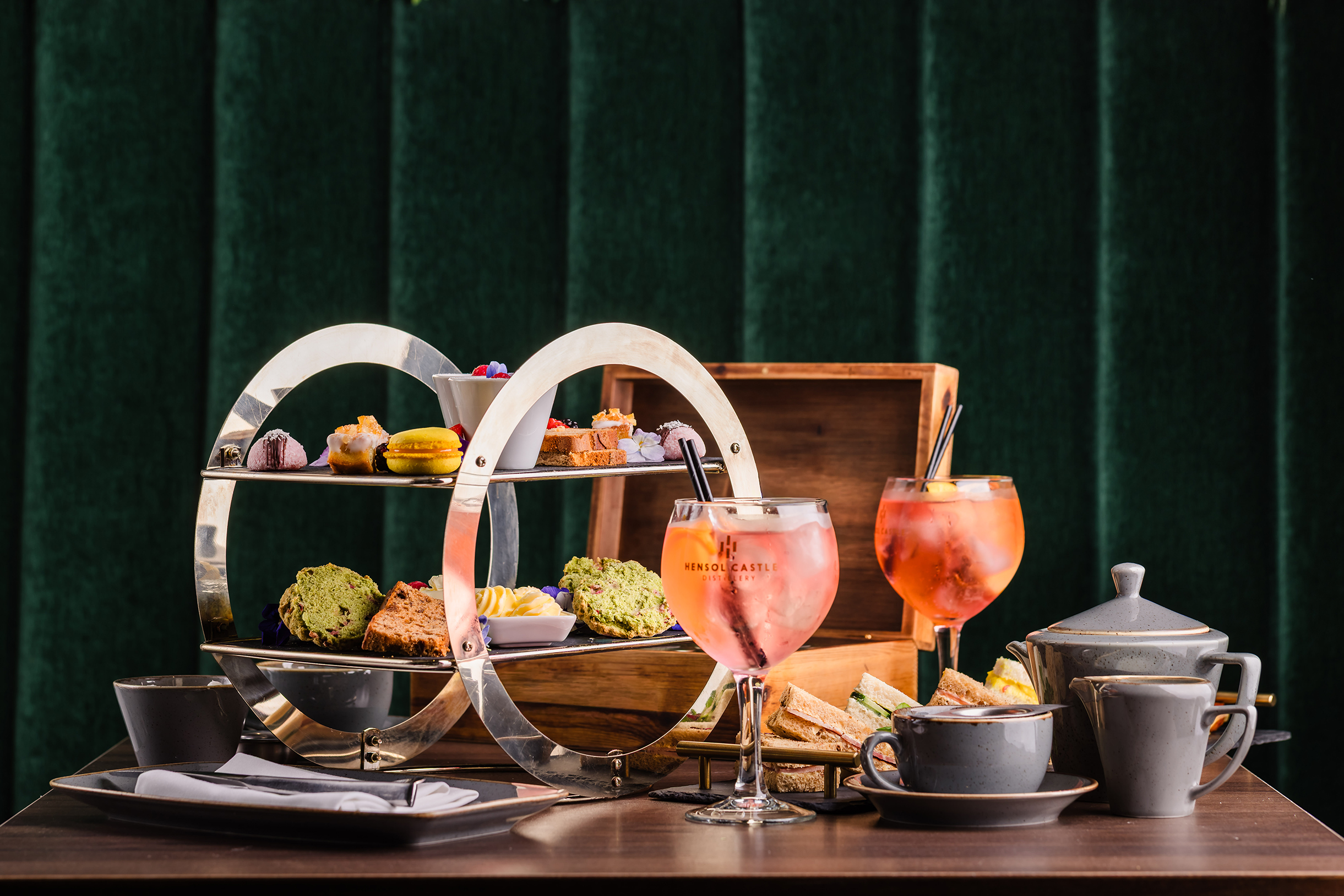 Afternoon tea set with pastries, sandwiches, tea, and pink drinks on a table against a green velvet backdrop.