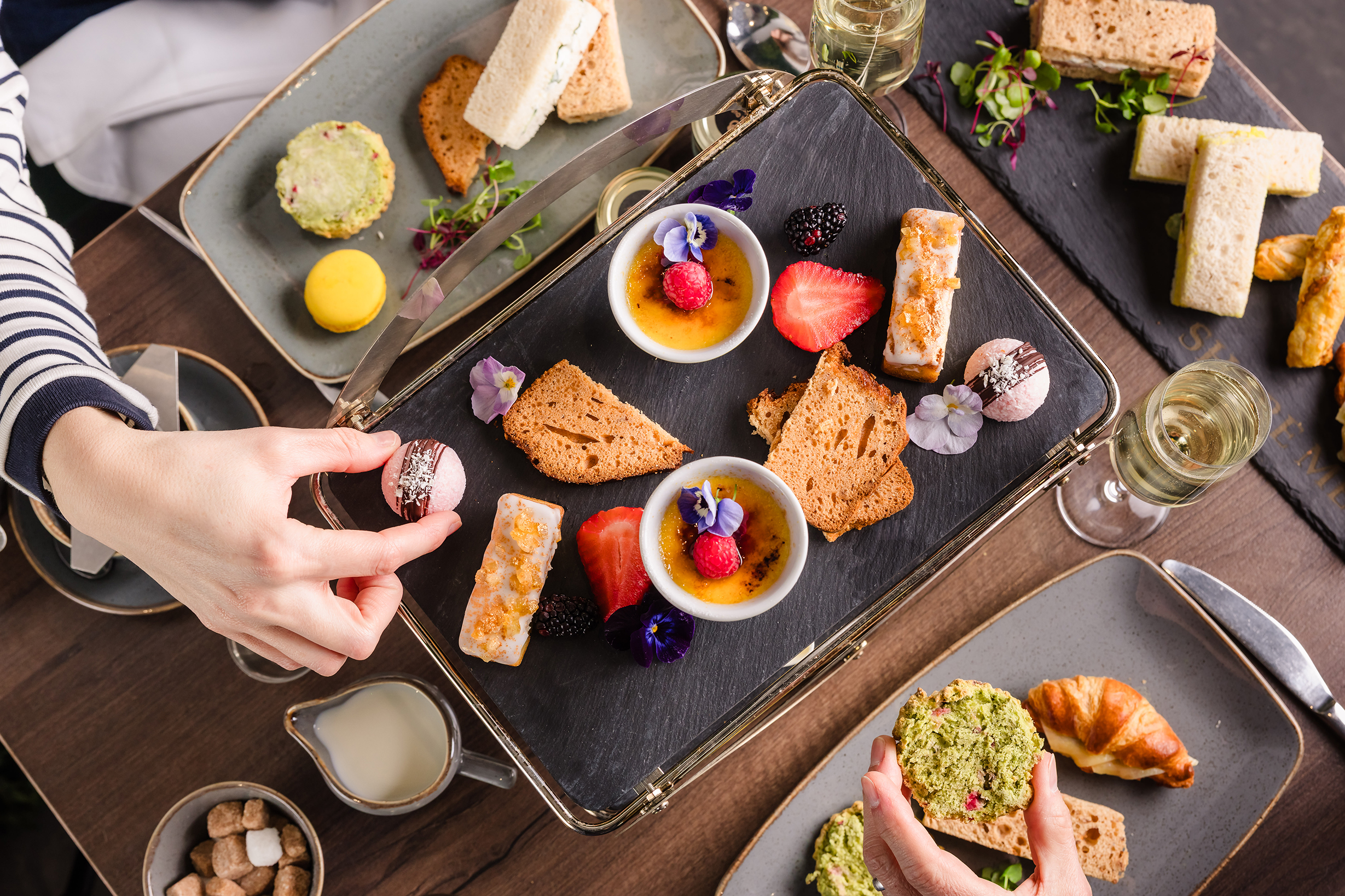 A table set for afternoon tea with assorted pastries, scones, fruit, and crème brûlée; hands reaching for treats.