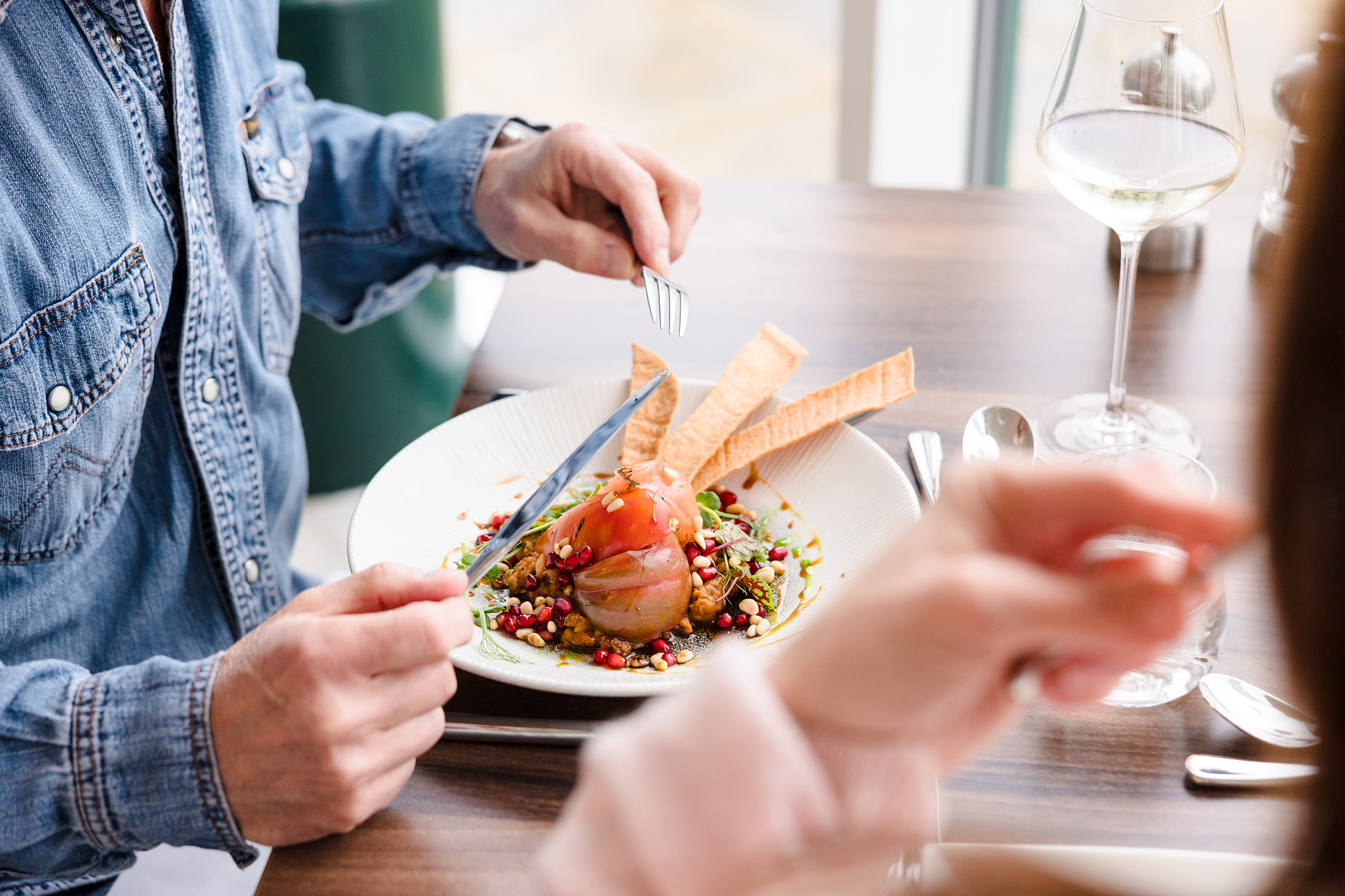 Person in a denim shirt enjoys sunday lunch with salmon, vegetables, pomegranate seeds, and bread crisps at a restaurant.