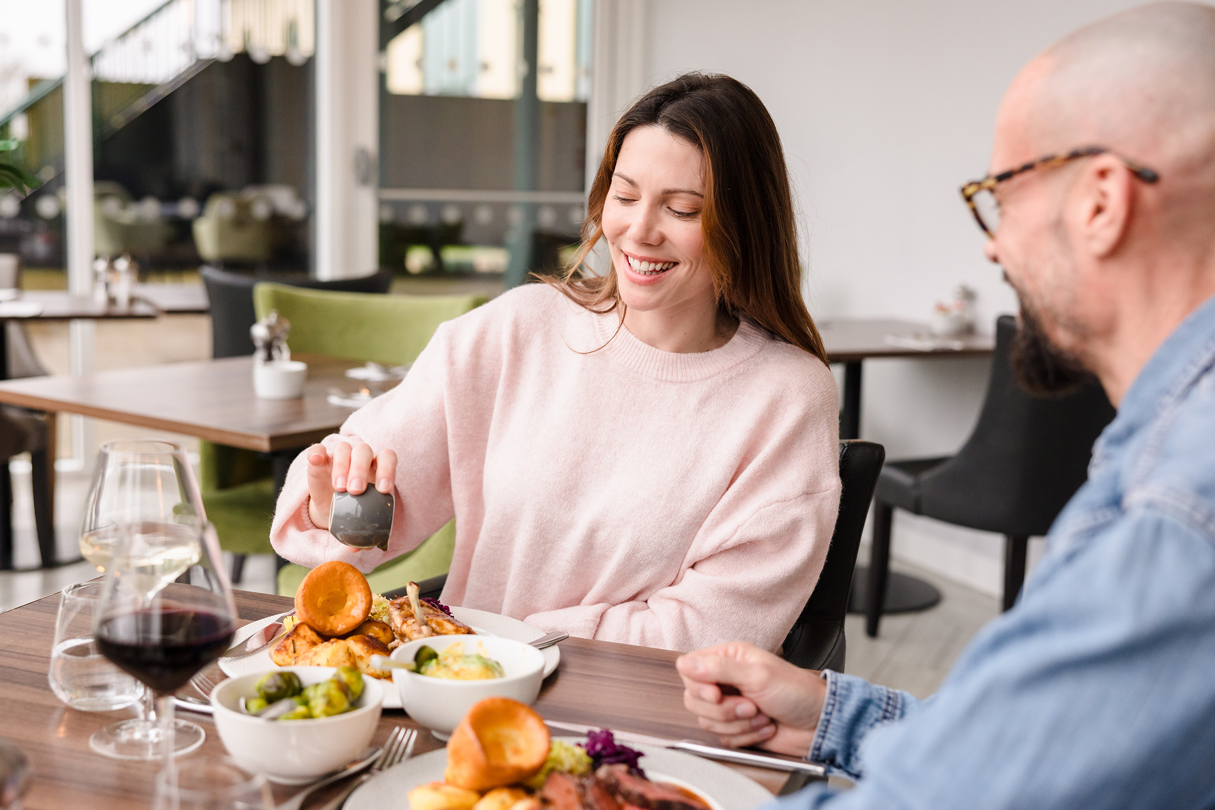A woman pours sauce on her meal while enjoying sunday lunch with a man at a restaurant table set with food and drinks.
