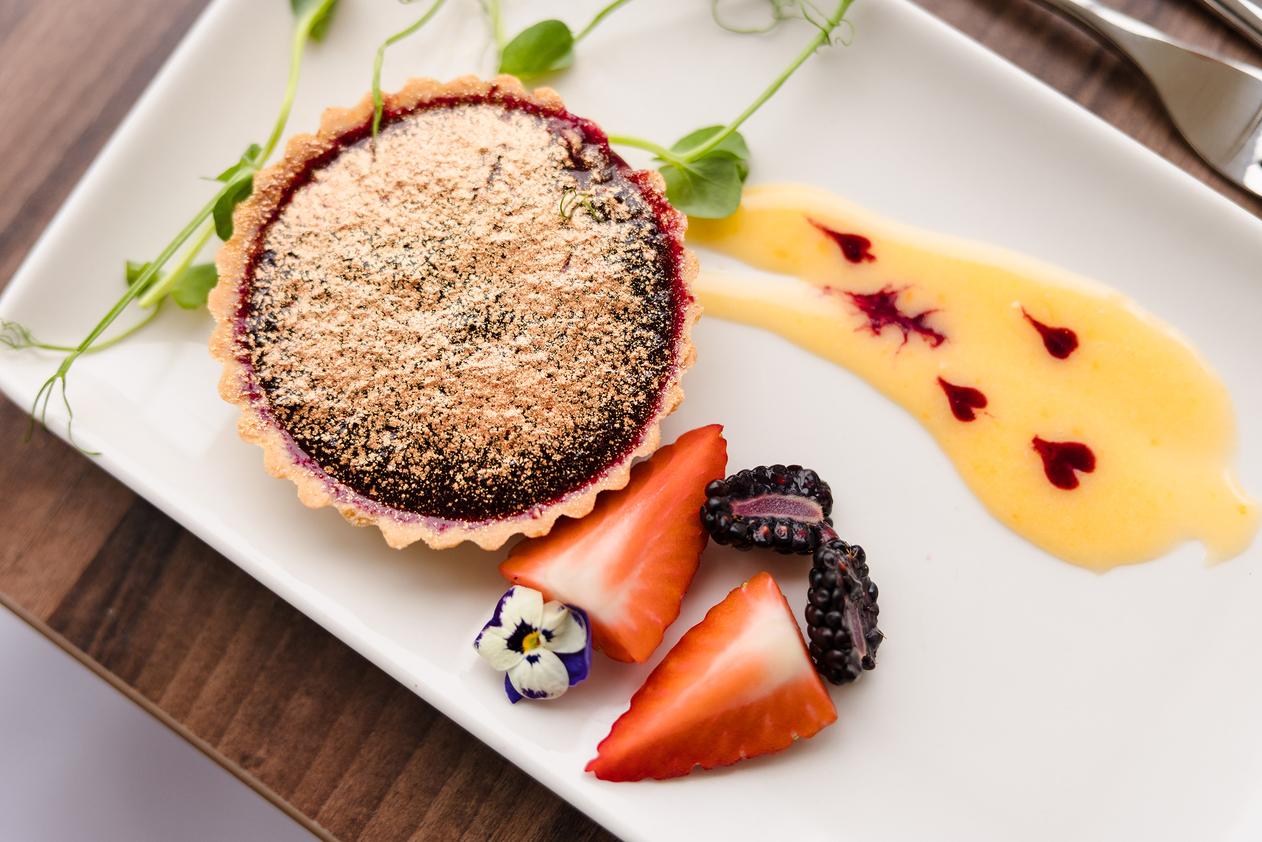 A fruit tart on a white plate, perfect for sunday lunch, with strawberries, blackberries, edible flowers, and microgreens.
