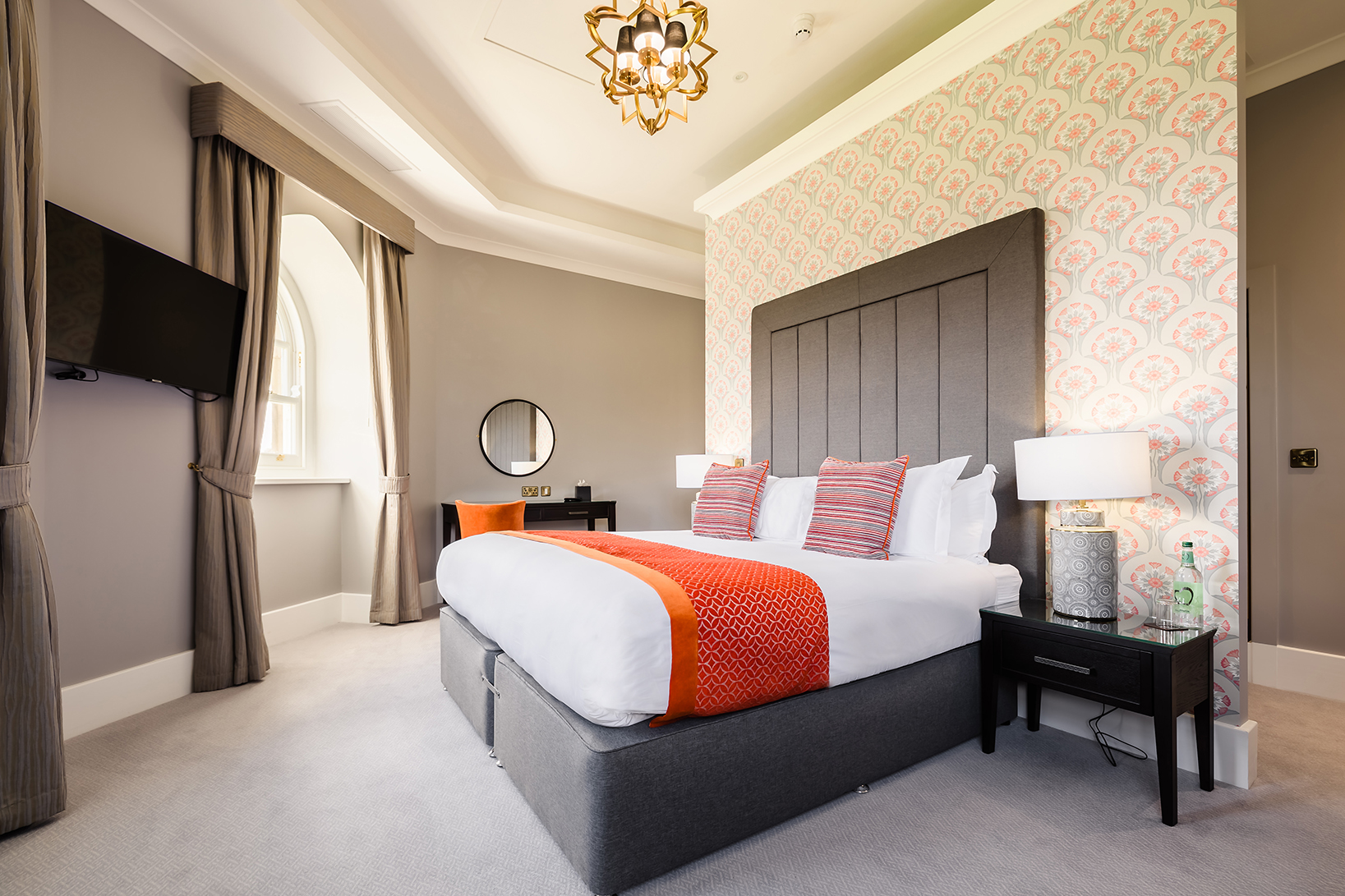Modern hotel room with a double bed, patterned accent wall, bedside tables, lamps, and a wall-mounted TV.