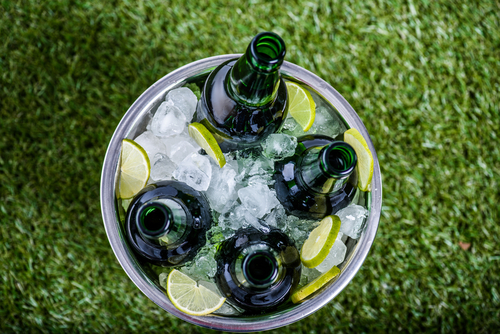 Five green bottles in a metal bucket with ice and lime slices, placed on green grass.