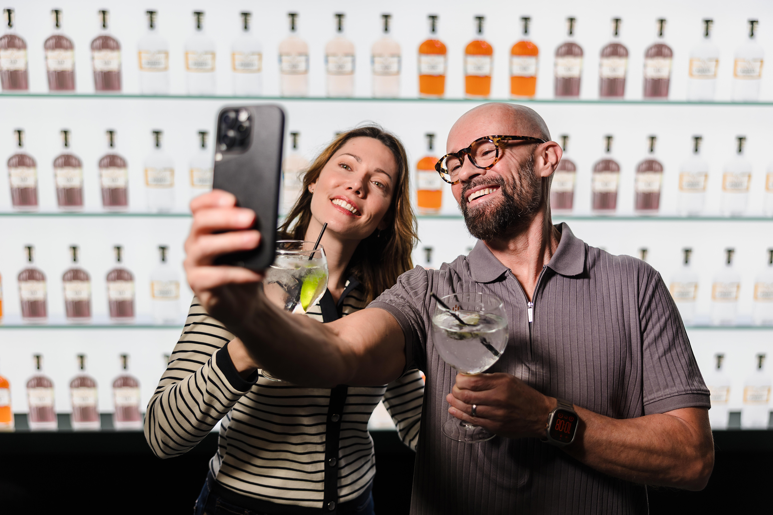 Two people smiling and taking a selfie with drinks, relaxing in luxury hotel rooms and suites, shelves of bottles behind them.