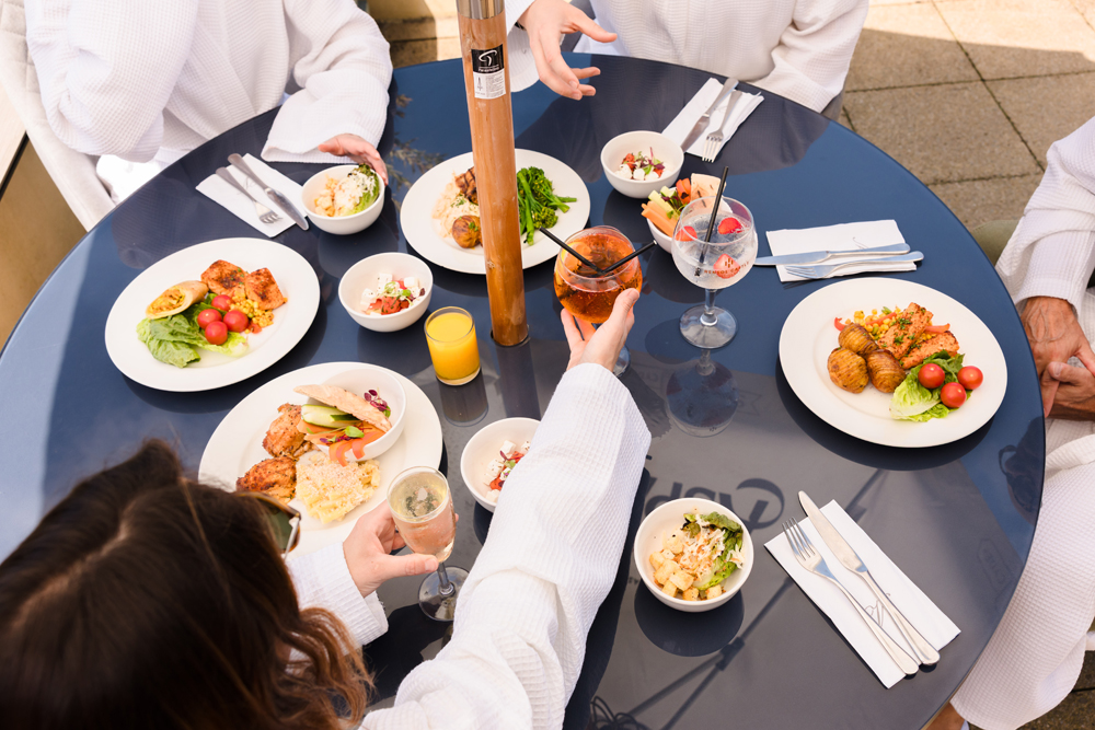 Four people in white robes enjoy a meal on a round outdoor table during relaxing Spa Breaks South Wales.