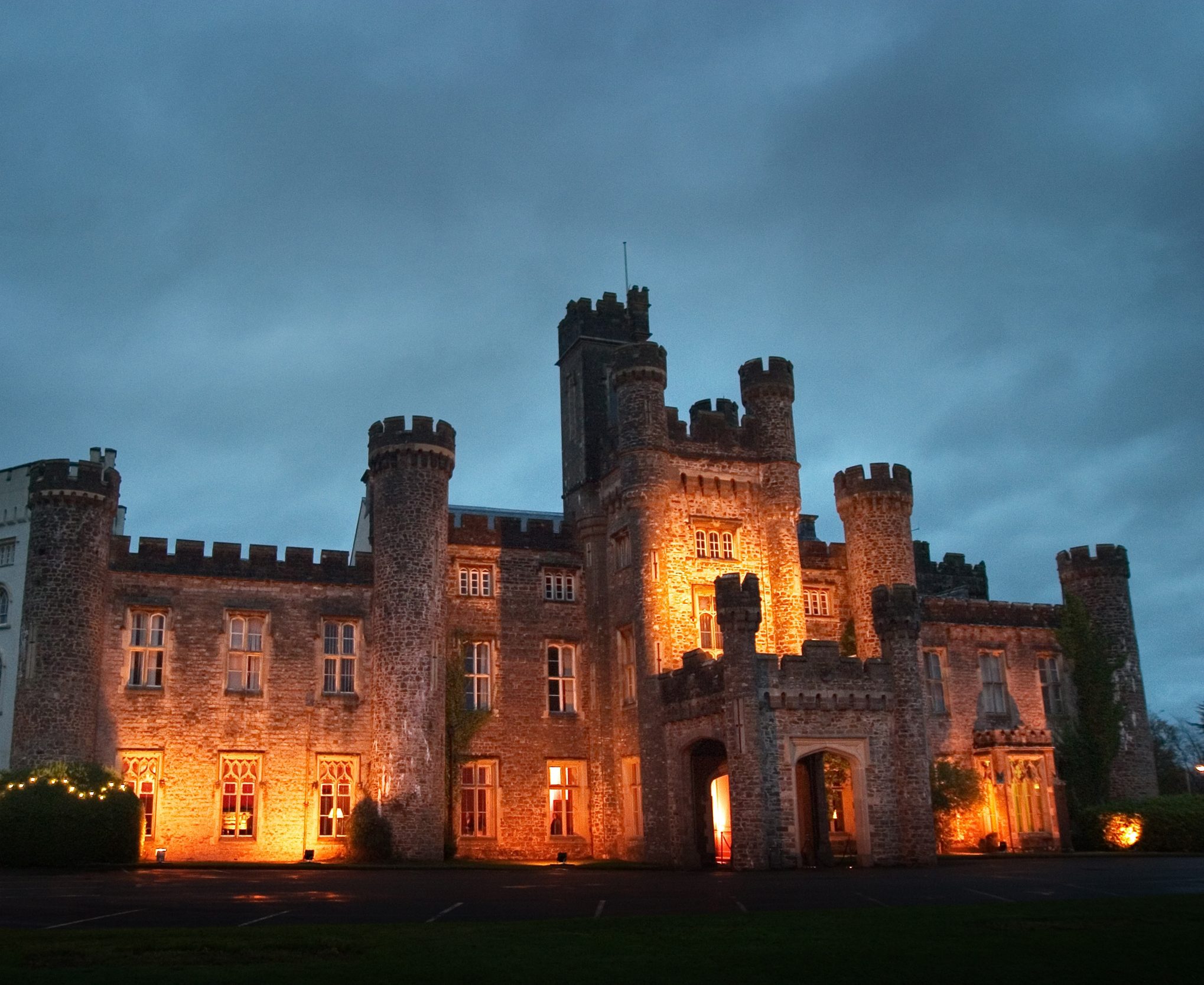 Stone castle illuminated by warm lights at dusk, perfect setting for a murder mystery, with tent on the lawn.