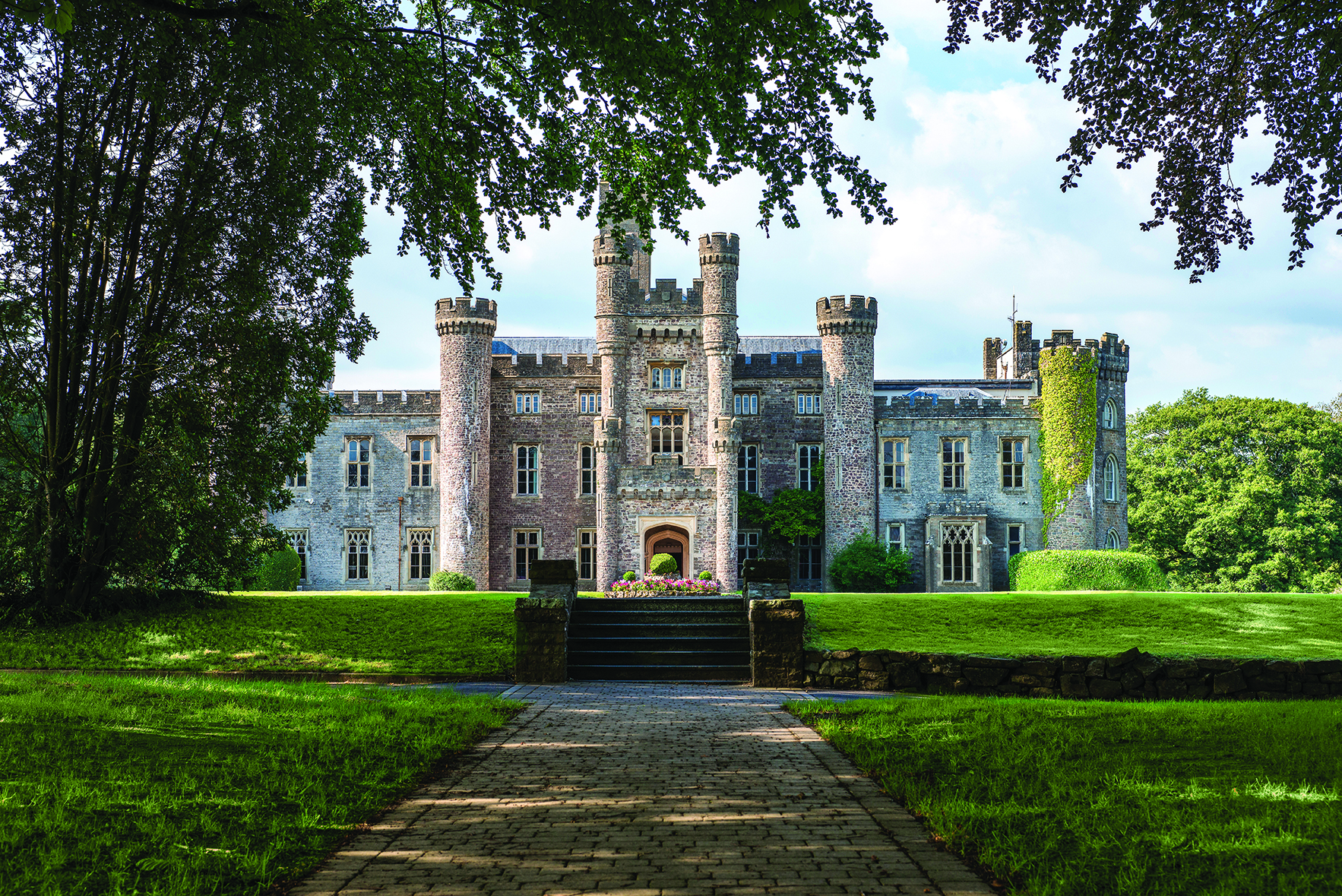 Large stone castle with multiple towers, set on a manicured lawn and surrounded by trees on a sunny day.