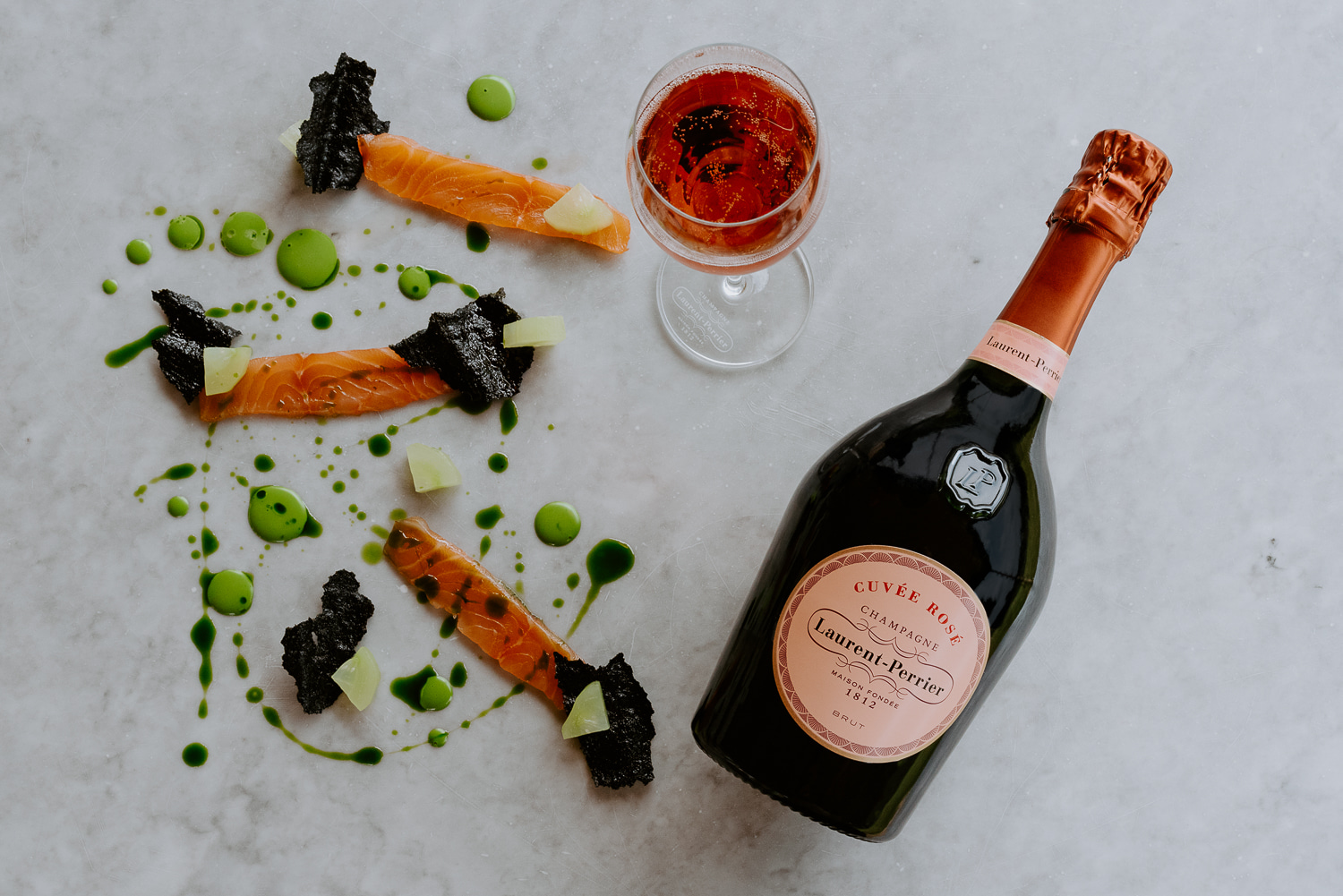 Bottle of Laurent-Perrier Cuvée Rosé, glass of rosé wine, and plated salmon with sauce and garnishes on a marble surface.